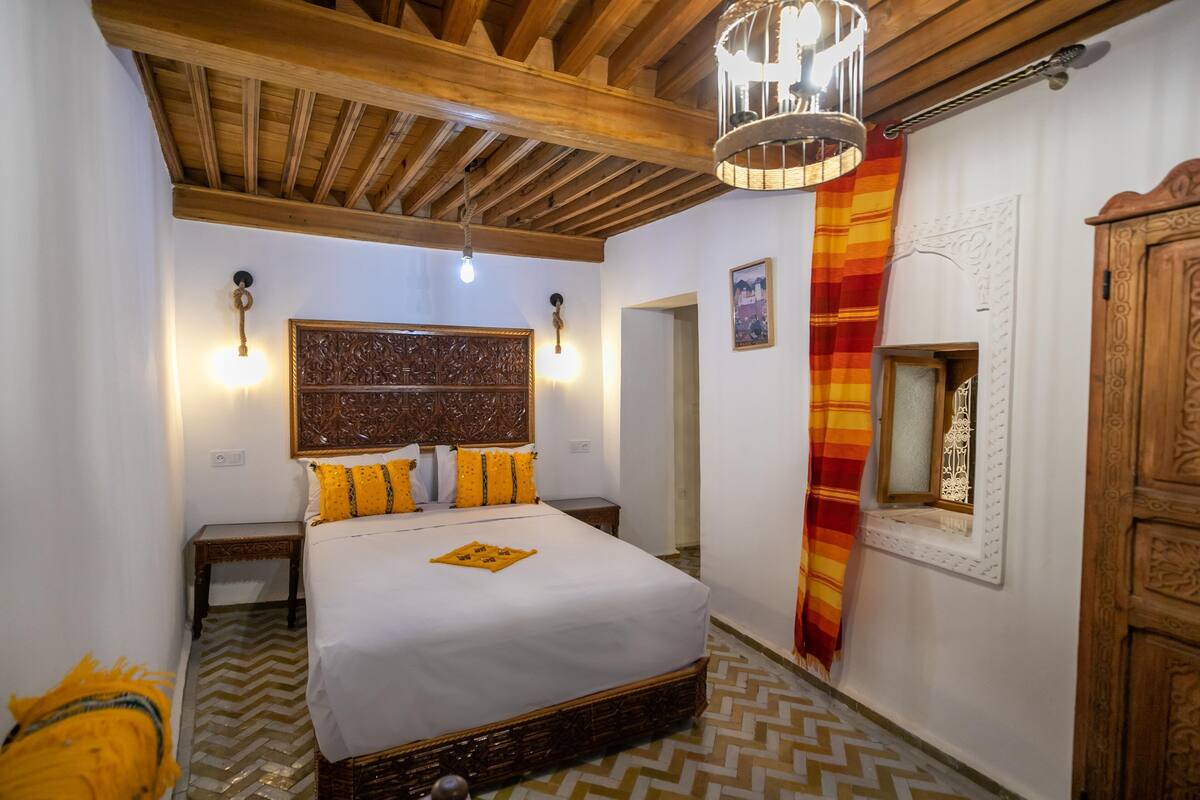 A bedroom with a large bed, wooden beams, a chandelier, and a patterned rug.