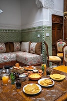 Uma área de refeições tradicional marroquina com uma mesa estampada, um sofá com estofamento floral e uma porta de madeira.