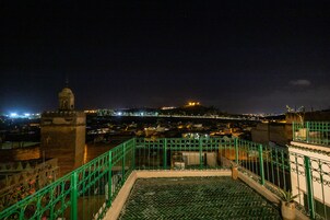 Vista noturna de um terraço no telhado, com guarda-corrimão verde, uma colina iluminada ao longe e um horizonte urbano ao fundo.