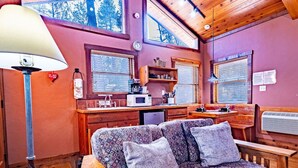 A cozy cabin interior with a sofa, a lamp, a kitchen area, and large windows. - Romantic Cabin | River Sounds | Hiking (Ruidoso)