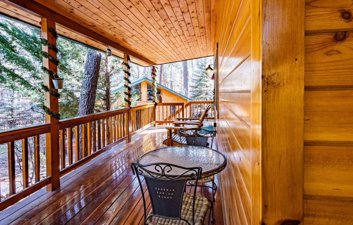 Une cabane en bois dotée d’une terrasse panoramique avec une table ronde et des chaises, entourée de rampes en bois et de grandes fenêtres.