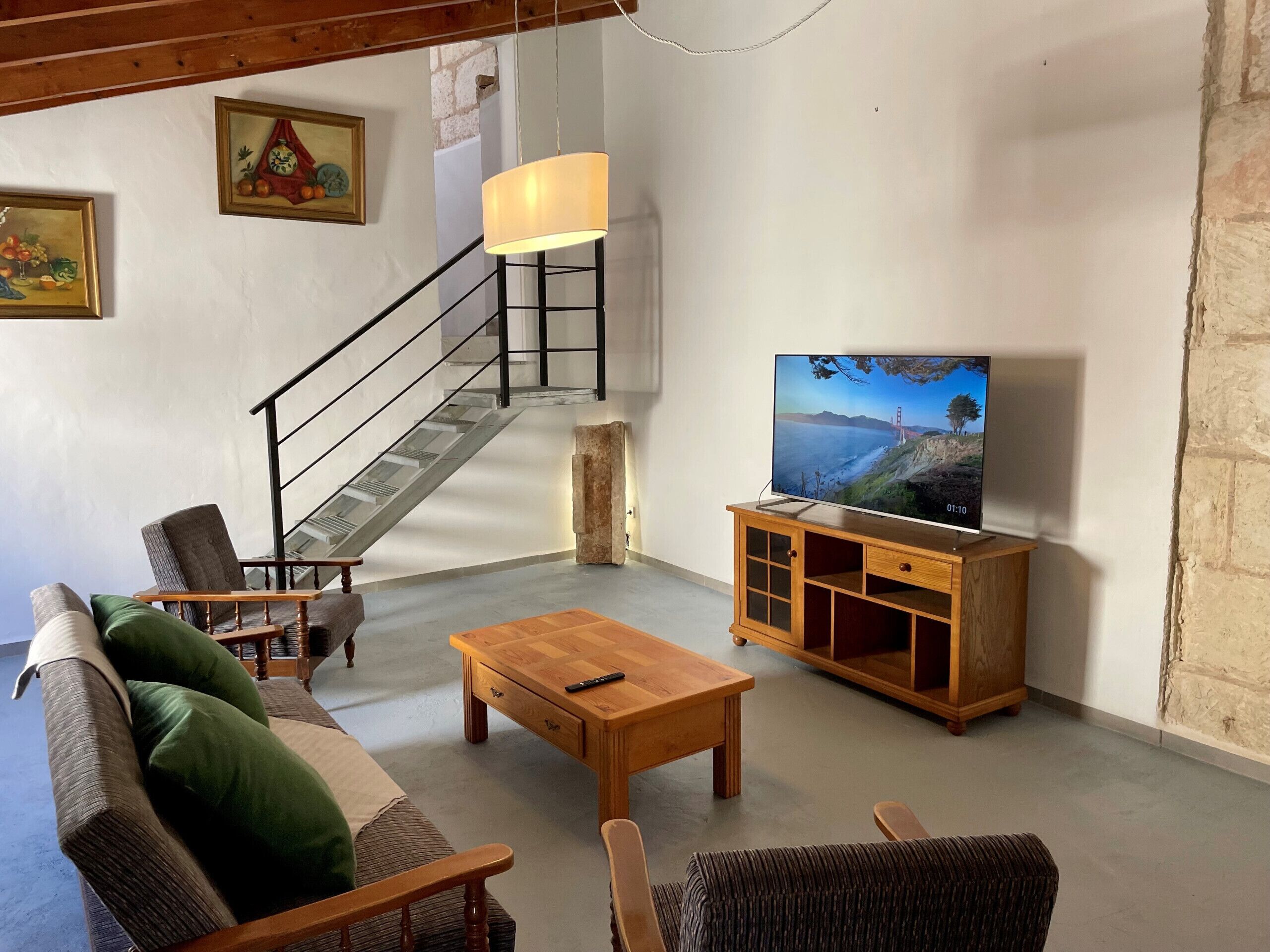 A living room with a sofa, chairs, a coffee table, a TV on a wooden cabinet, a staircase, and a wall-mounted lamp.