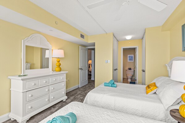 Une chambre d’hôtel avec deux lits, une commode blanche, un miroir et une salle de bain visible à travers une porte ouverte.