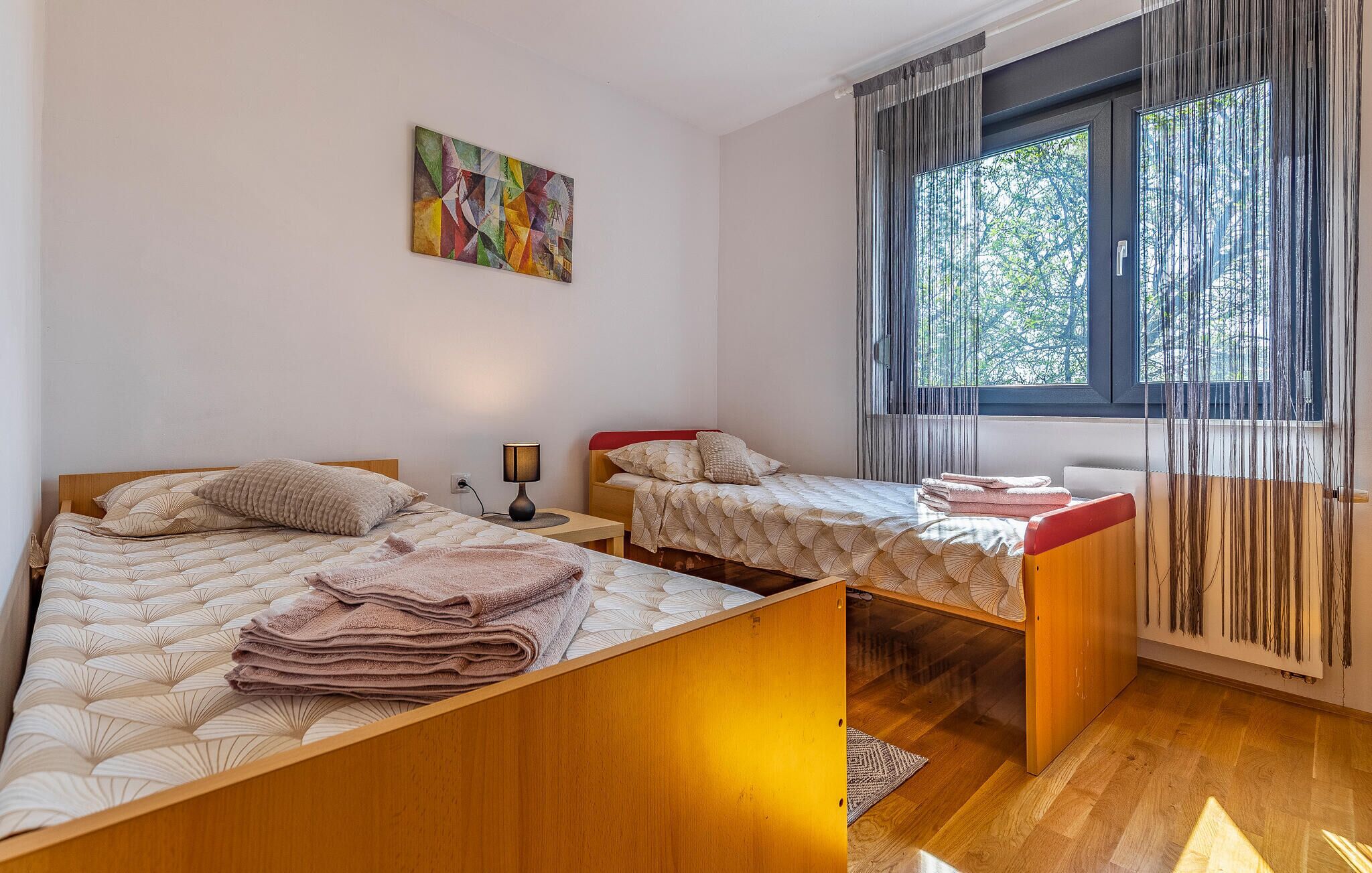 Une chambre avec deux lits, une table de chevet avec une lampe, un tableau au mur et une fenêtre avec des rideaux.
