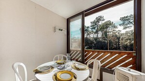 Une table de salle à manger ronde, mise avec des assiettes, des couverts et des verres, entourée de chaises blanches, et offrant une vue sur les arbres à travers une grande fenêtre.
