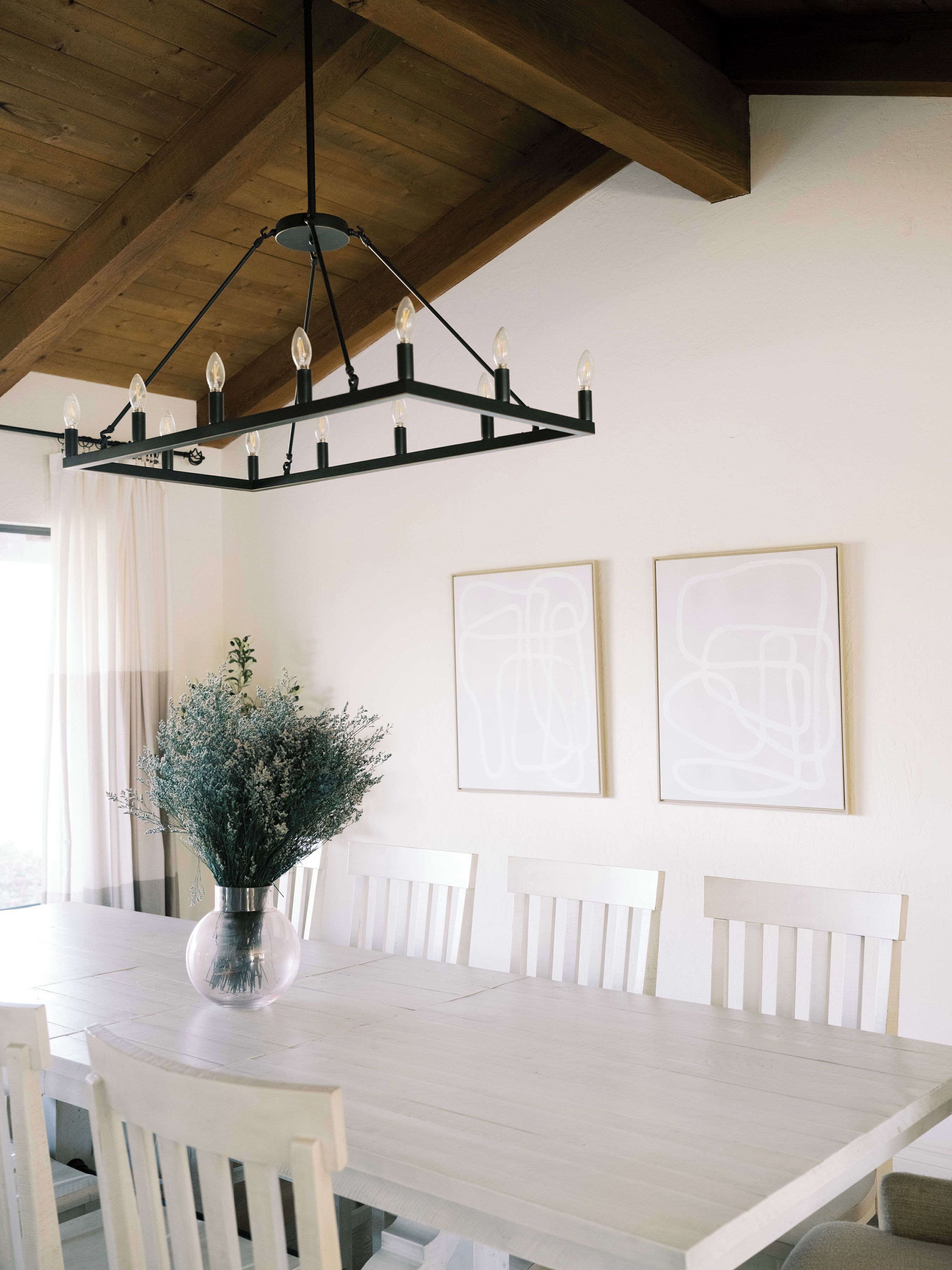A dining area with a wooden ceiling, a chandelier, a white dining table, and two framed abstract artworks on the wall.