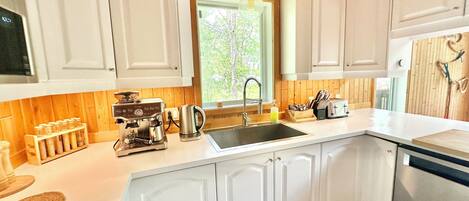 Cocina con armarios blancos, una cafetera, un lavabo y una ventana con vistas a árboles.
