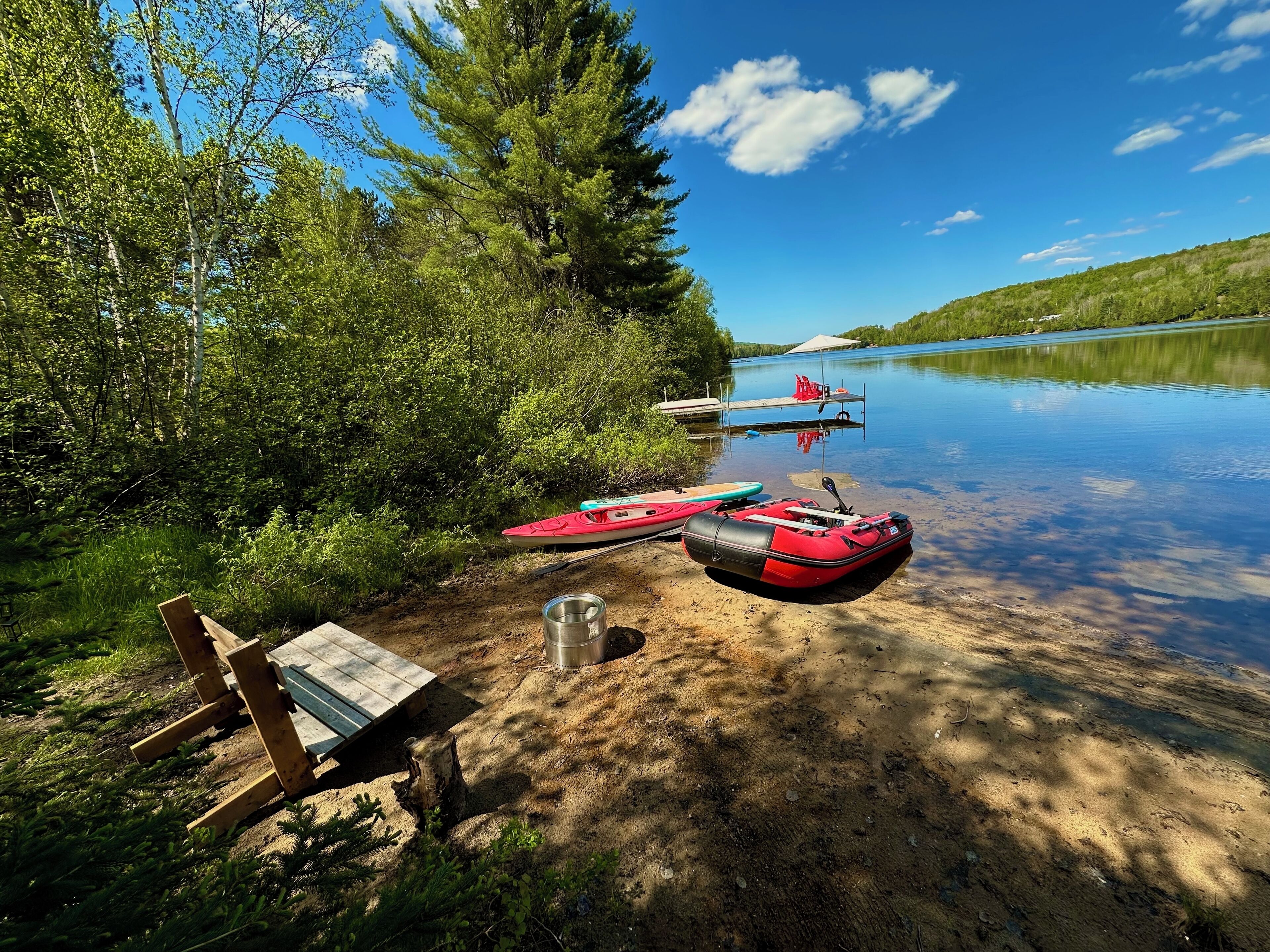 Zona de camping junto al lago, con un banco de madera, una taza de metal y un bote hinchable rojo.