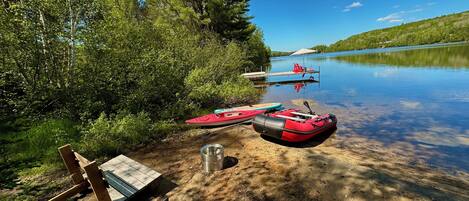 Zona de camping junto al lago, con un banco de madera, una taza de metal y un bote hinchable rojo.
