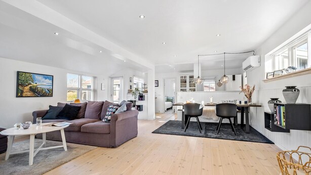 A spacious living area with a sofa, coffee table, and dining table. A kitchen is visible in the background with modern appliances and pendant lighting. - 3 bedroom stunning home in Højby (Højby)