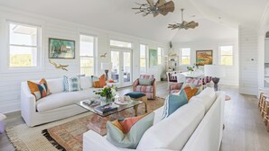 A spacious living room with white walls, large windows, and a variety of seating options including a white sofa and a patterned armchair. - Chic Shellter, Bald Head Island, NC (Bald Head Island)