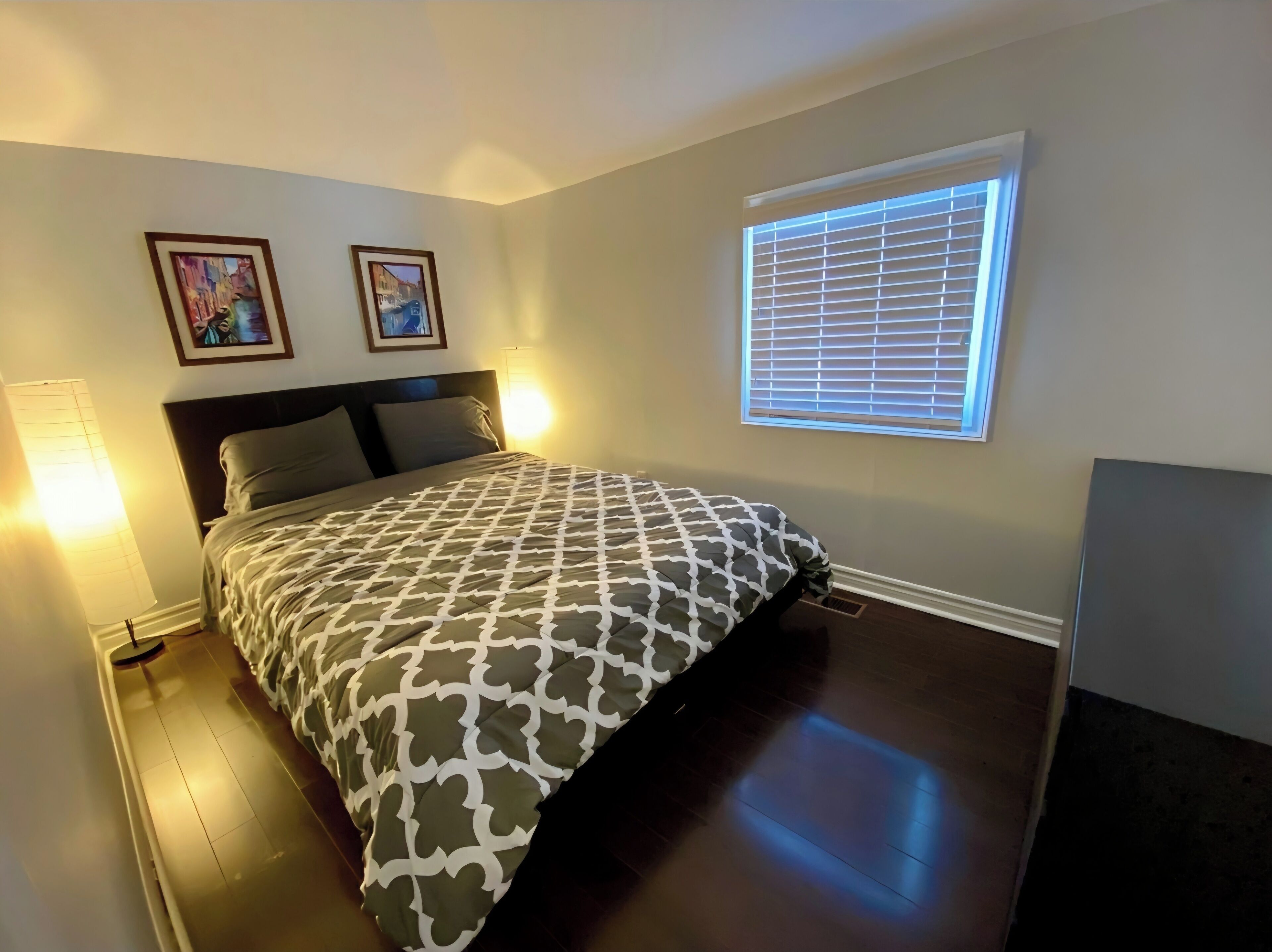 A bedroom with a bed, two wall-mounted lamps, a window with blinds, and two framed pictures on the wall.