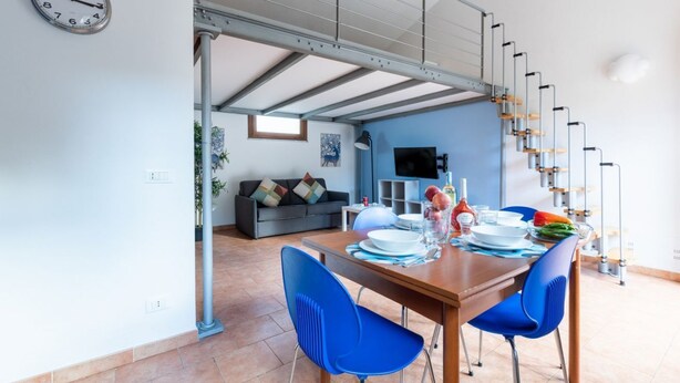 A dining area with a table set for two, a sofa in the background, and a staircase leading to an upper level. - GuestHost - Cozy studio apartment of about 50 sqm with mezzanine, located on the ground floor (one step at the entrance), able to comfortably accommodate up to 4 people. The accommodation is located in a quiet neighborhood, with all the main services. Wi (Pisa)