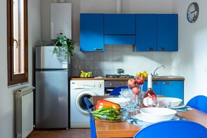 A compact kitchen with blue cabinets, a refrigerator, a washing machine, and a dining area with a table set for two.