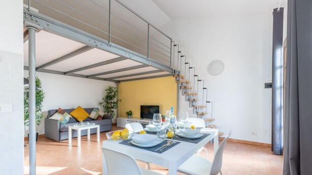A dining area with a table set for four, a sofa, and a staircase leading to an upper level. - GuestHost - Cozy studio apartment of about 50 sqm with mezzanine, located on the ground floor (one step at the entrance), able to comfortably accommodate up to 4 people. The accommodation is located in a quiet neighborhood, with all the main services. Wi (Pisa)