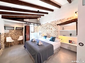 A bedroom with a stone wall, a bed with blue pillows, a wooden ceiling, and a small shelf with books and a plant.