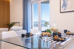 Quarto de hotel moderno com mesa de jantar de vidro posta para o café da manhã, cama com roupa de cama branca e vista para o mar e montanhas por grandes janelas.