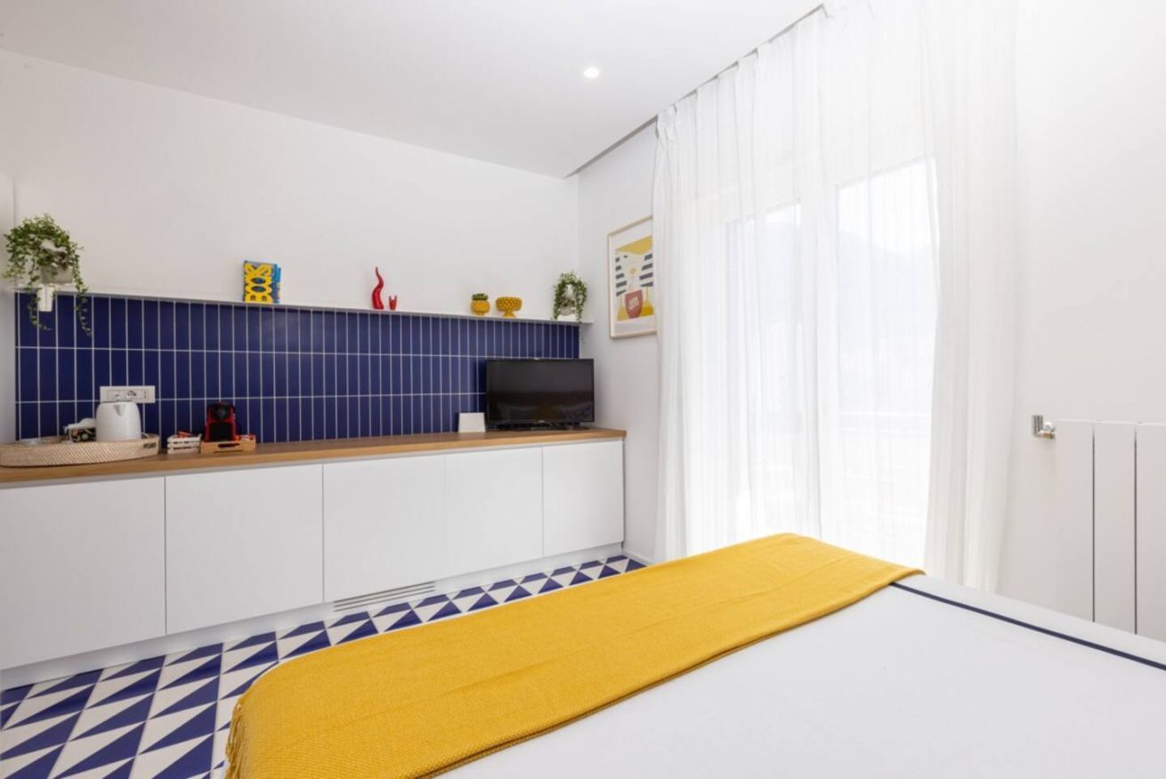 Een moderne hotelkamer met een kitchenette, een bed met een gele deken en een groot raam met witte gordijnen.
