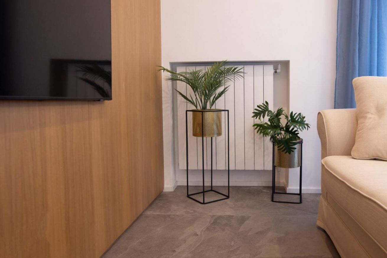 En moderne stue med et fladskærms-tv, to høje gulvlamper med planter, en beige sofa og en radiator.