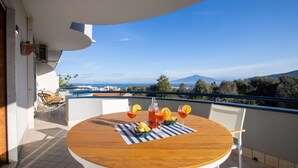 Une table ronde en bois, recouverte d’une nappe à rayures, deux chaises blanches et une bouteille de boisson rouge agrémentée de rondelles de citron, le tout sur un balcon offrant une vue dégagée sur la mer et les montagnes.