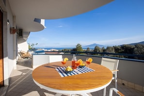 A round wooden table with a striped tablecloth, two white chairs, and a bottle of red drink with lemon slices on a balcony with a clear view of the sea and mountains.