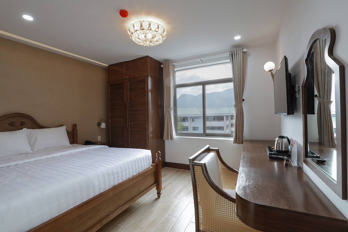 A hotel room with a bed, a wooden wardrobe, a desk with a kettle, a chair, a large mirror, and a view of mountains through the window.