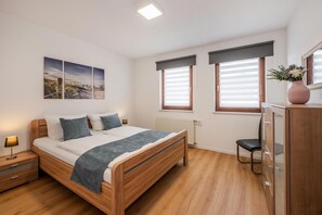 Ein Schlafzimmer mit einem hölzernen Bett, Nachttischen, einem Schreibtisch und einem Sessel. Zwei Fenster mit Jalousien, ein Heizkörper und ein an der Wand befestigtes Kunstwerk sind ebenfalls vorhanden.