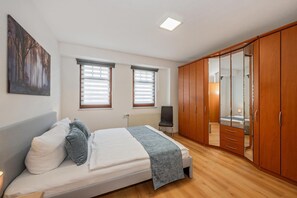 A bedroom with a bed, two windows with blinds, a radiator, a wardrobe, and a mirror.