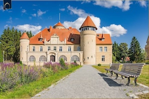 A castle-like building with a central tower, arched windows, and a paved pathway leading to a bench.