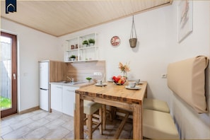 A compact kitchen with a refrigerator, sink, and wooden cabinets. There is a dining table with chairs and a small shelf above it.