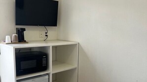 A small, compact room with a bed, a white shelving unit, a microwave, and a television. - Rogue River Landing #31A (Gold Beach)