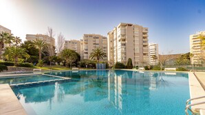 Une piscine à l’eau d’un bleu cristallin, entourée de palmiers et bordée d’une rangée d’immeubles à plusieurs étages dotés de balcons.