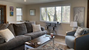 A living room with a gray sofa, a coffee table, and a window with a view of trees. - Cozy creekside home with deck, 15 min to airport, shopping, 1 hr to Crater Lk. 
 (Eagle Point)