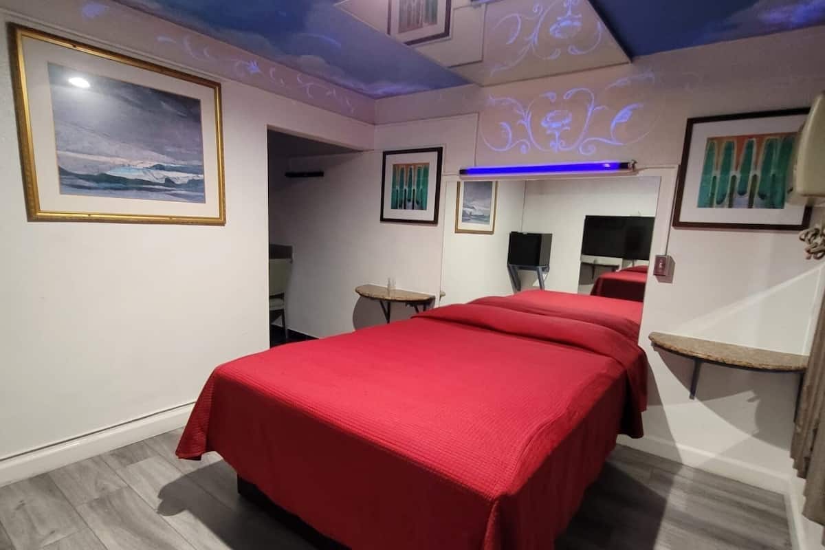 A bedroom with a large bed, a desk, and a chair. There are framed pictures on the walls and a ceiling fan.