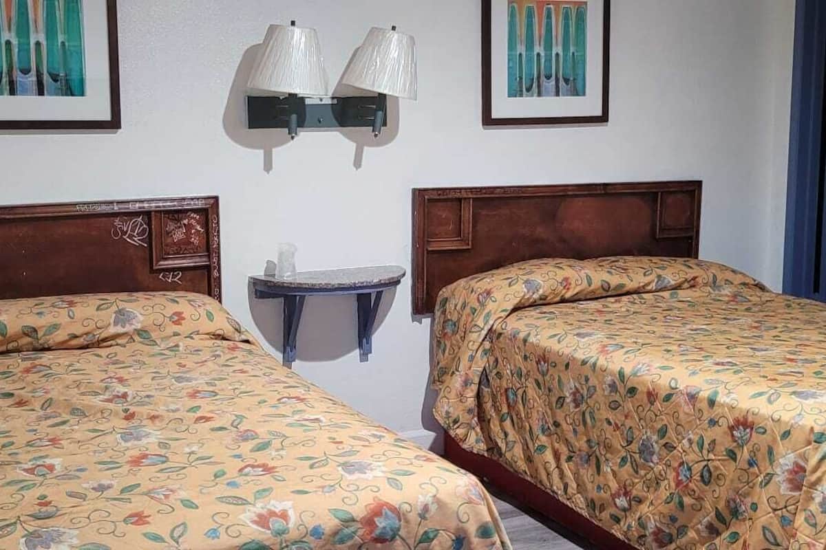 A room with two wooden beds with floral bedspreads, a small round table, and two wall-mounted lamps.