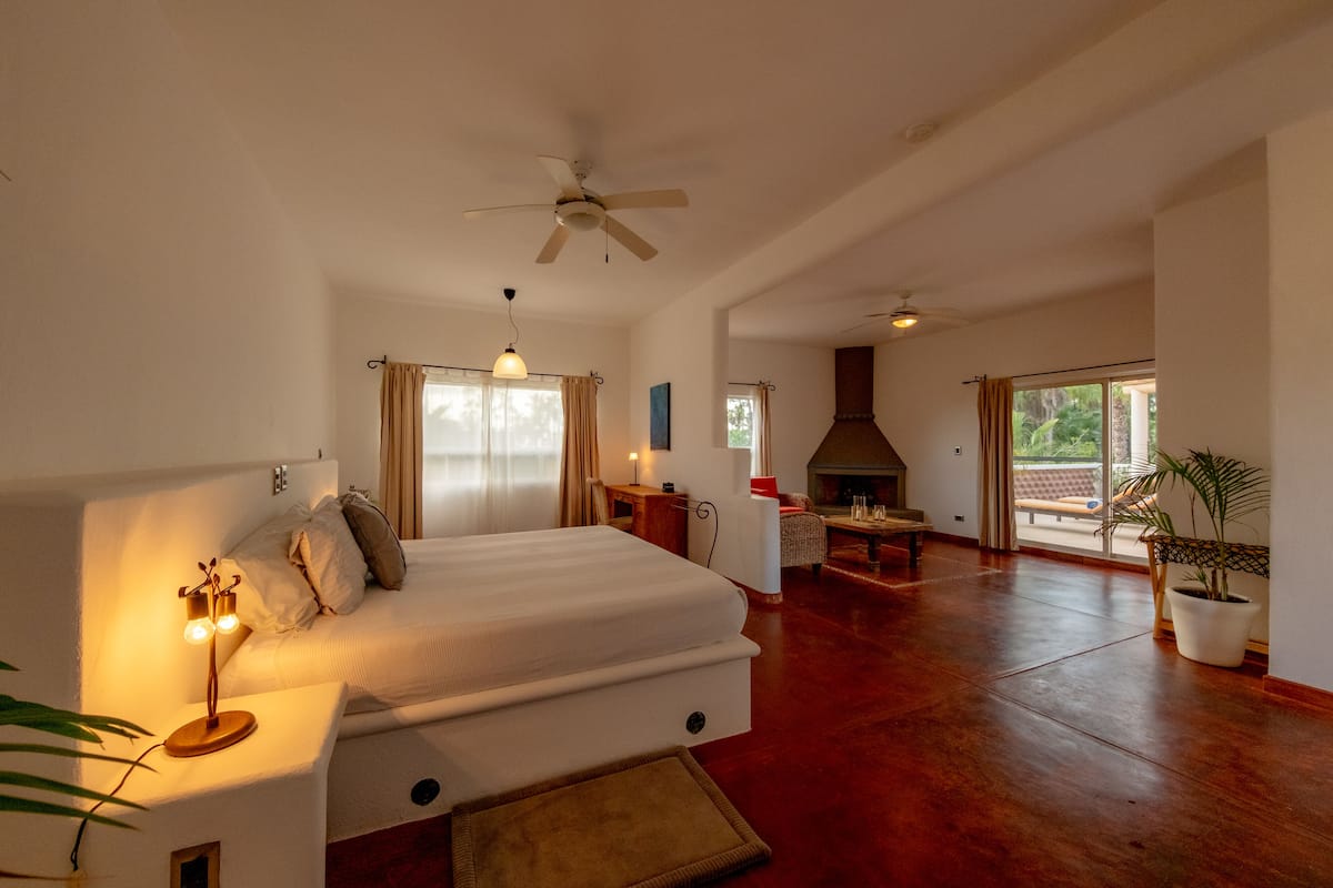 A spacious bedroom with a large bed, a ceiling fan, a fireplace, and a view of the outdoors.