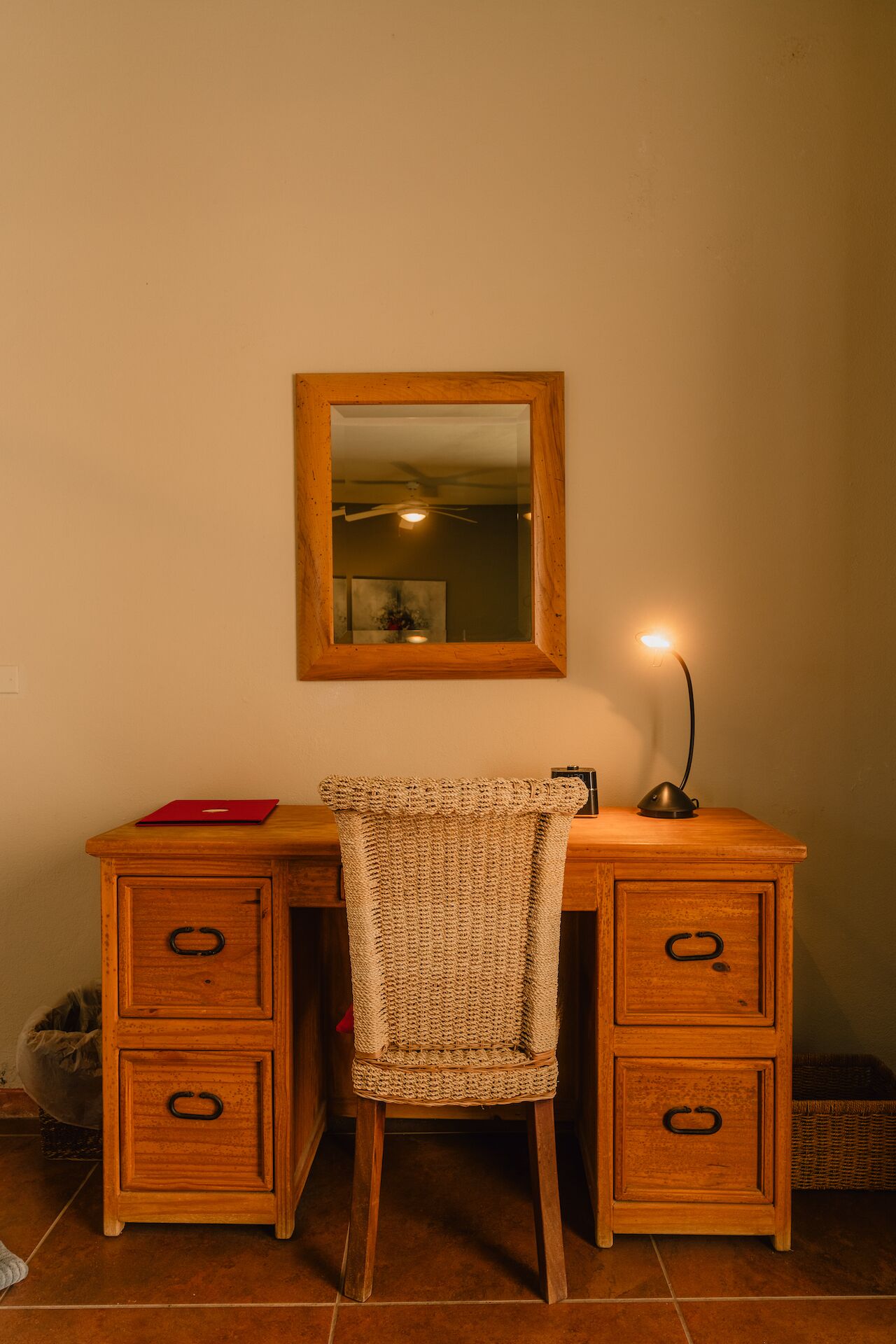 A wooden desk with a woven chair, a mirror, and a desk lamp.