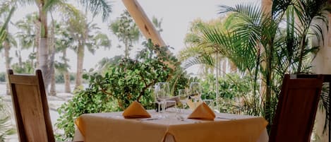 A table set for two with a yellow tablecloth, two wooden chairs, and a tropical backdrop with palm trees.