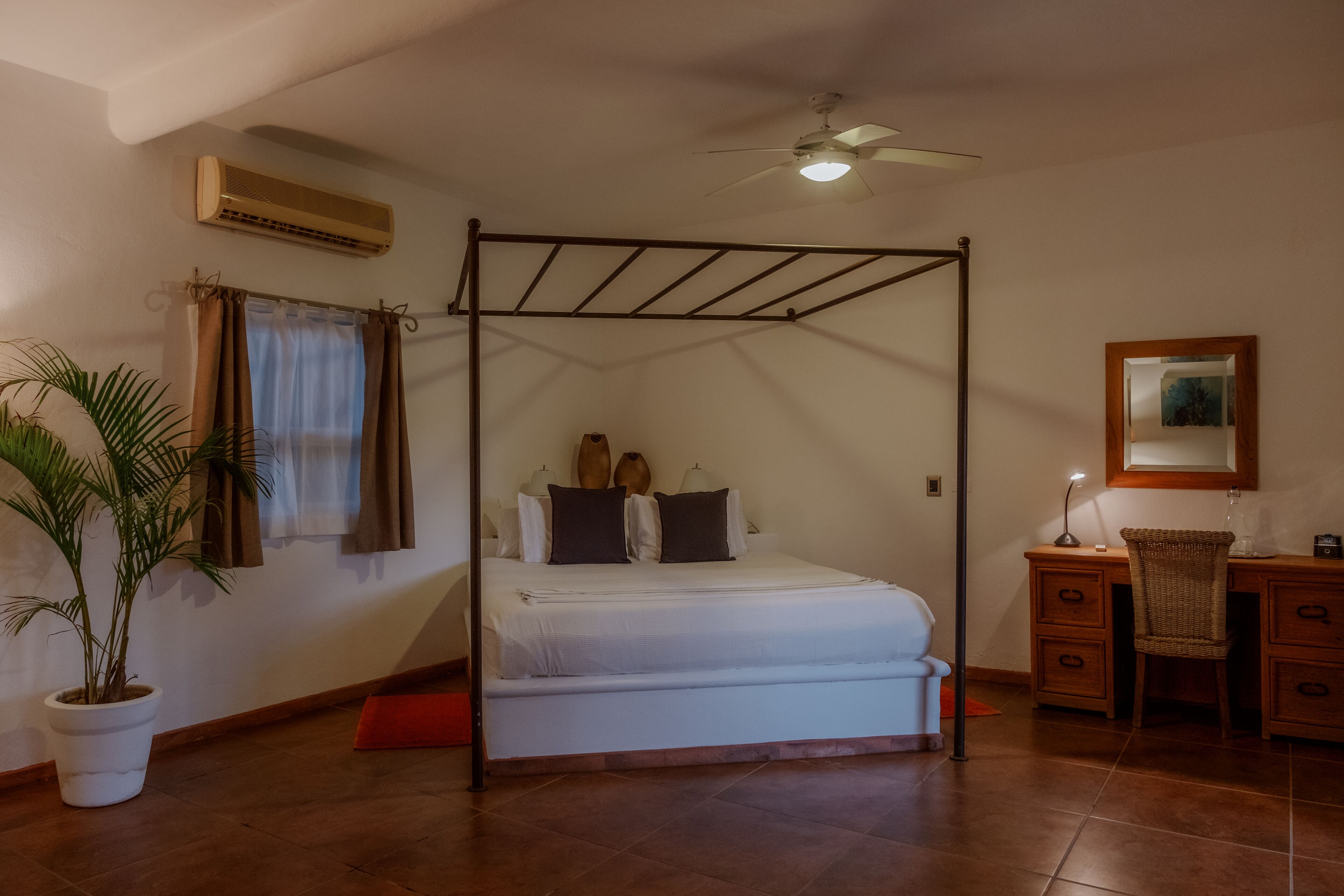 Una cama con dosel, un ventilador de techo, un escritorio con cajones, una silla, una planta en maceta y un cuadro en la pared.