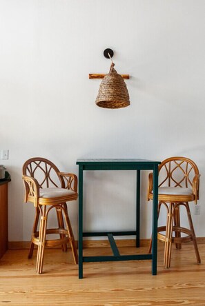 Una habitación con piso de madera, dos sillas de mimbre, una mesa verde y una lámpara colgante de fibras.