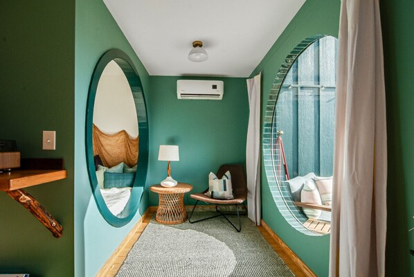 Una habitación con paredes color turquesa, un espejo grande, una mesita redonda con una lámpara, un sillón de mimbre y una ventana con cortinas.