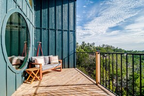 Un balcón con piso de madera, una zona de estar con cojines y vista a zonas verdes.