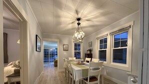 Ein Essbereich mit einem Tisch für vier Personen, weißen Stühlen, einer Chandelierleuchtre und großen Fenstern mit Vorhängen.