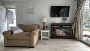 Ein Wohnzimmer mit einem beigen Sofa, einem kleinen Beistelltisch mit Lampe, einem Kamin und einem an der Wand befestigten Fernseher.