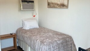 Un dormitorio pequeño y ordenado con una cama individual, una mesita de noche, una silla y un aire acondicionado instalado en la pared.
