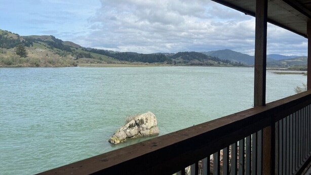 A view from a covered area overlooking a calm lake with a rocky outcrop in the water, surrounded by hills. - Studio Suite #38 River Front (Gold Beach)