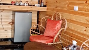 A cozy wooden cabin interior with a round wooden table, wicker chairs, a small refrigerator, and a shelf with jars and a kettle. - orchid huts (Ajloun)