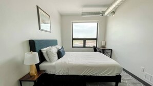A bedroom with a bed, a nightstand with a lamp, a window with a view, and a wall-mounted air vent. - Sleepover | Alluring 1BD/1BA - Des Moines (West Des Moines)
