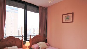 A hotel room with two beds, a nightstand with a lamp, a framed picture on the wall, and a view of tall buildings through the window. - Beautiful apartment for 4 guests with pool, WIFI, A/C, TV and pets allowed (Benidorm)
