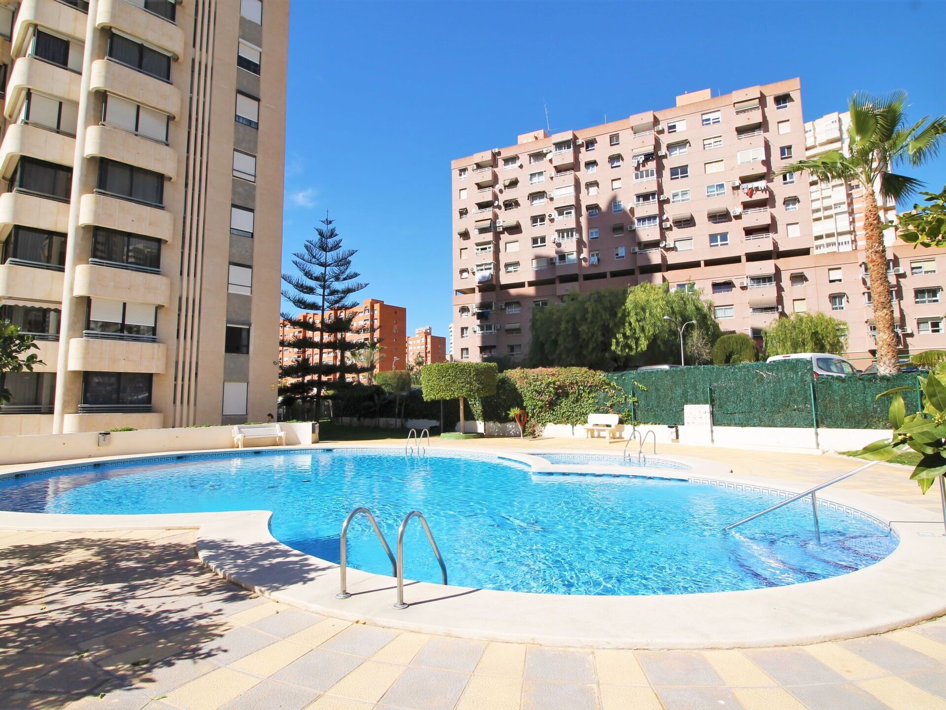 Ein Pool mit kreisförmigem Design, umgeben von einer gepflasterten Fläche mit Stufen zum Pool hin. Im Hintergrund sind mehrstöckige Wohngebäude mit Balkonen und eine hohe Palme zu sehen.